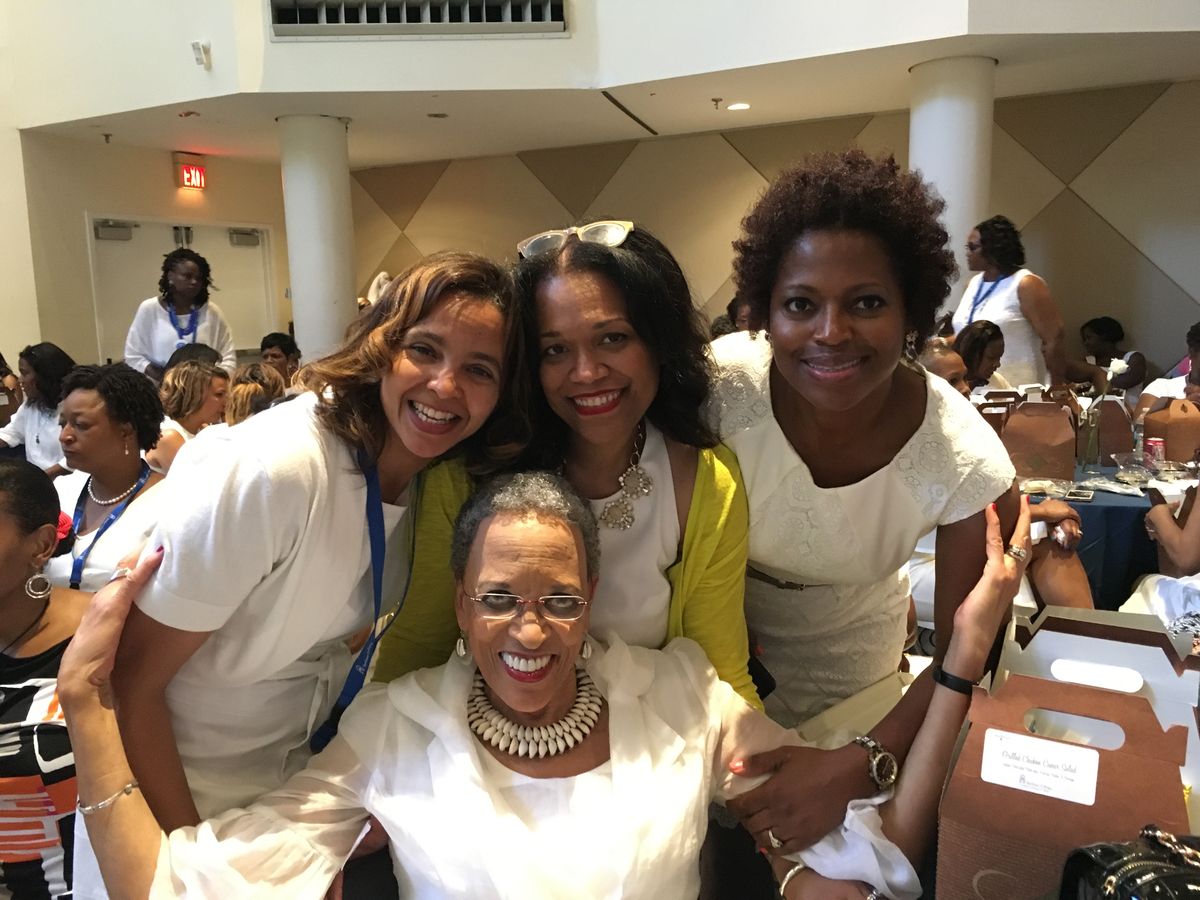 Four women dressed in white smiling together at a crowded luncheon